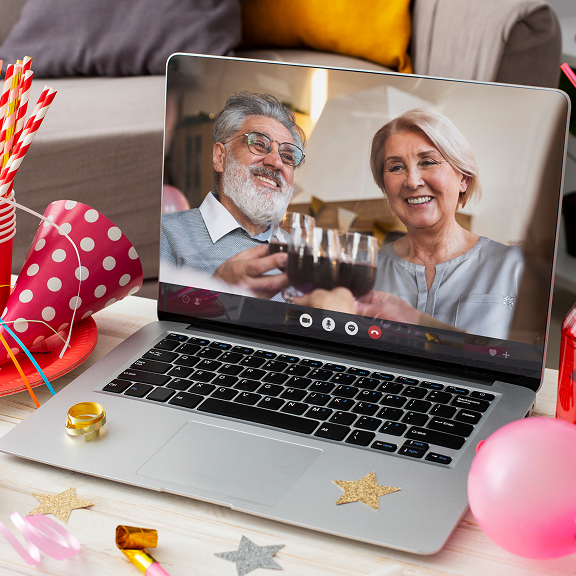 Laptop showing a smiling elderly couple clinking glasses on a video call, surrounded by party decorations including balloons and a crown.