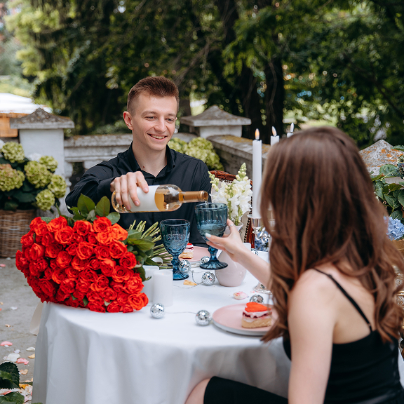 Man pouring wine into a glass held by a woman at an outdoor table decorated with red roses and candles.