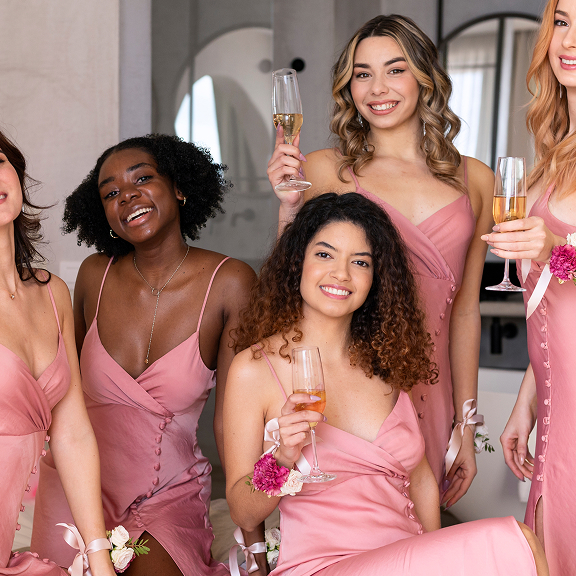 Group of smiling bridesmaids in pink dresses holding champagne glasses and floral wrist corsages.