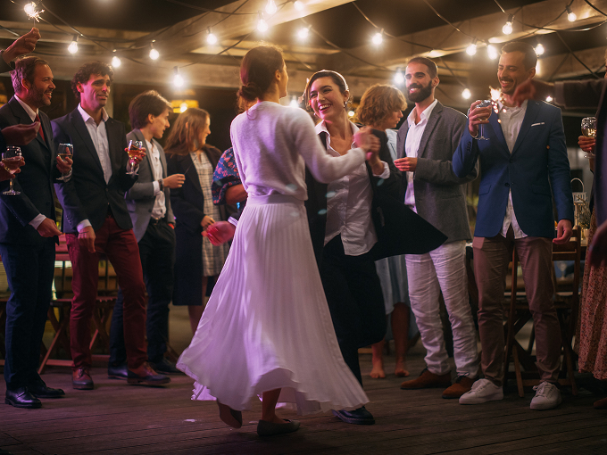 A group of people dressed in formal attire celebrating at an outdoor wedding, with bride and guest dancing in the center and others holding drinks and sparklers under string lights.