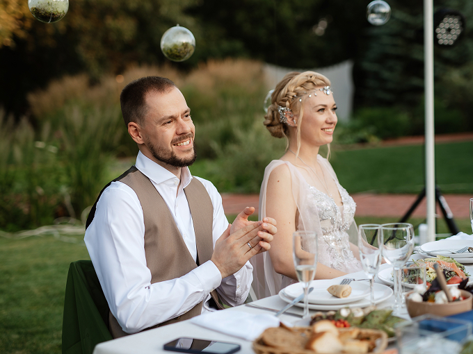 Bride and groom smiling and sitting at an outdoor wedding reception table with food and glassware.