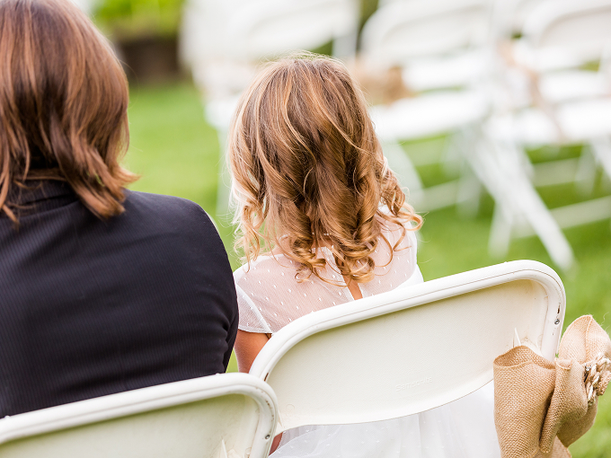 A woman and a young girl with curled hair sitting on white folding chairs during wedding ceremony.