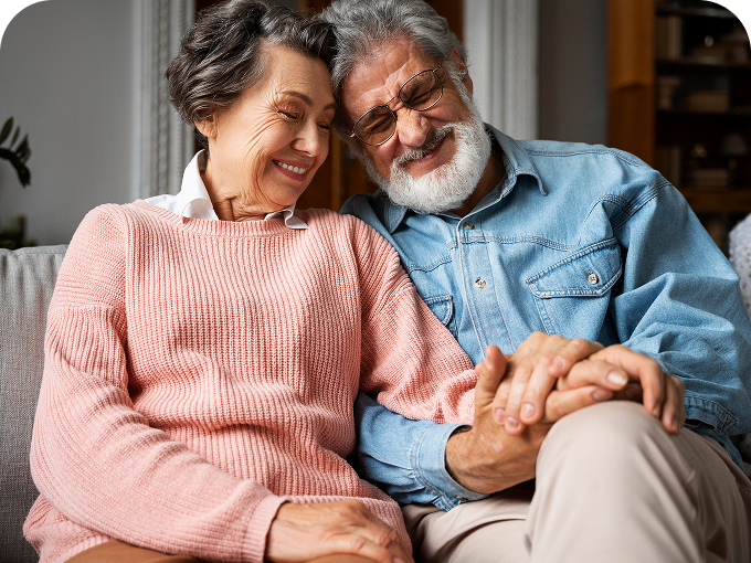 Smiling elderly couple sitting close on a couch, holding hands and leaning on each other.