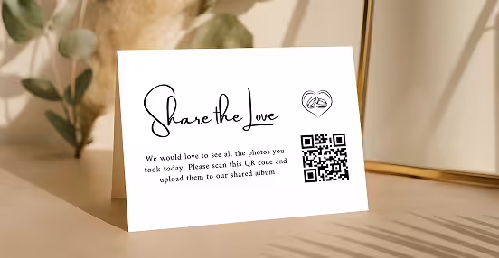 A white card on a wooden surface with text 'Share the love,' a small heart illustration with two hands, a QR code, and a message asking to scan and upload photos to a shared album.