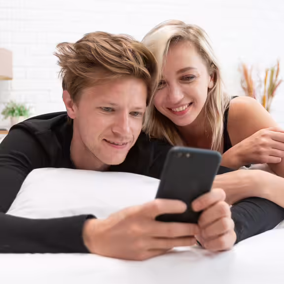 Smiling young couple lying on a bed looking at a smartphone together.