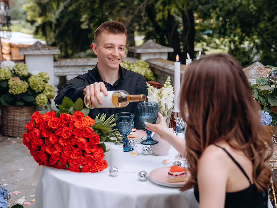Young man smiling while pouring a drink into a blue glass held by a woman sitting at an outdoor table decorated with a large bouquet of red roses and candles.