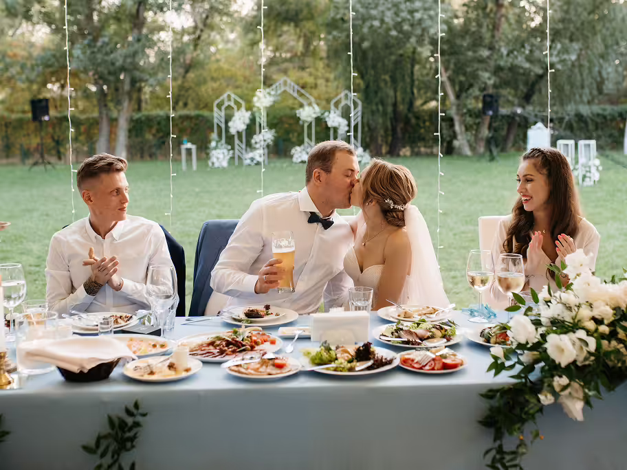 Bride and groom kissing at a decorated wedding reception table while guests on both sides clap and smile.