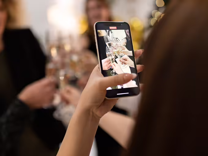 Close-up of a person holding a smartphone capturing a photo of people toasting with champagne glasses at a celebration.
