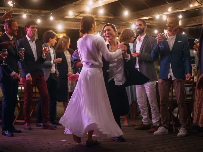Two women dancing joyfully at a party with guests around holding drinks under string lights.