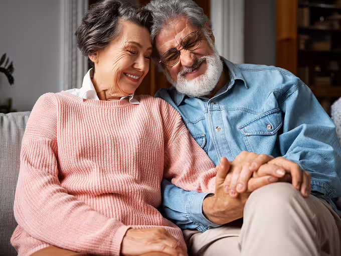 Elderly couple sitting closely on a couch, holding hands and smiling with their eyes closed.