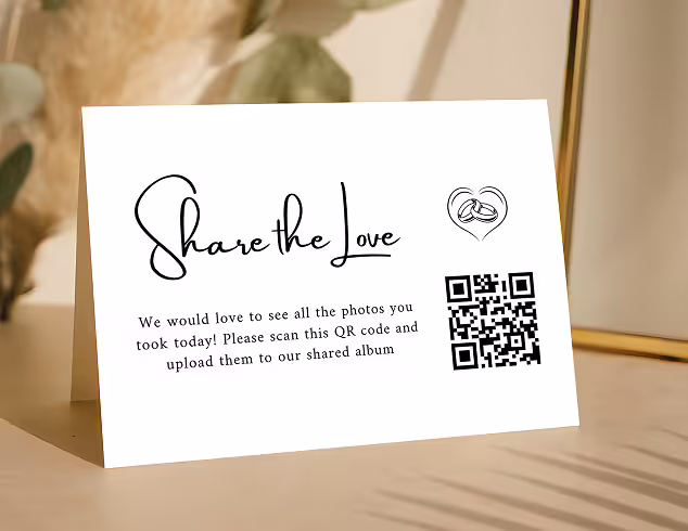 Table tent card with text 'Share the Love' requesting guests to scan a QR code to upload photos to a shared album, featuring a heart with wedding rings icon.