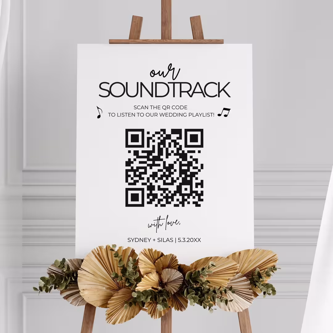 White sign on wooden easel with a QR code and text inviting guests to scan for a wedding playlist, decorated with dried palm leaves and eucalyptus.
