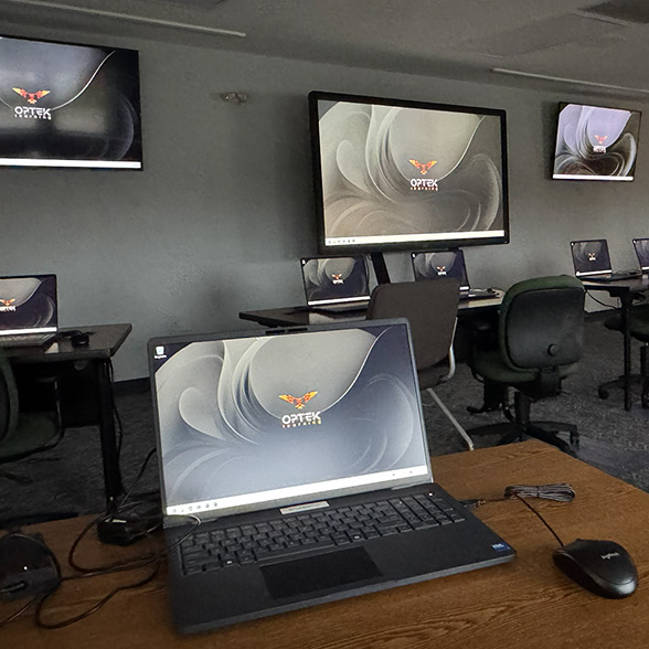 Laptop on a wooden desk with multiple screens displaying the Optek Systems logo in a classroom or training room.