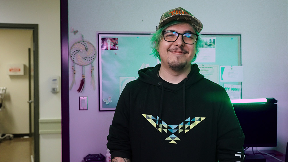 Smiling person with green hair wearing a patterned cap, glasses, and a black hoodie with a geometric design, standing indoors near a bulletin board.