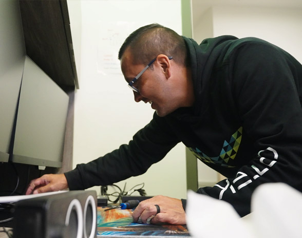 Man wearing glasses and a black hoodie adjusting equipment under a desk in an office.