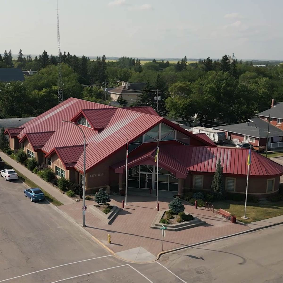 A red-roofed city hall building with three flagpoles, surrounded by trees and neighboring houses.