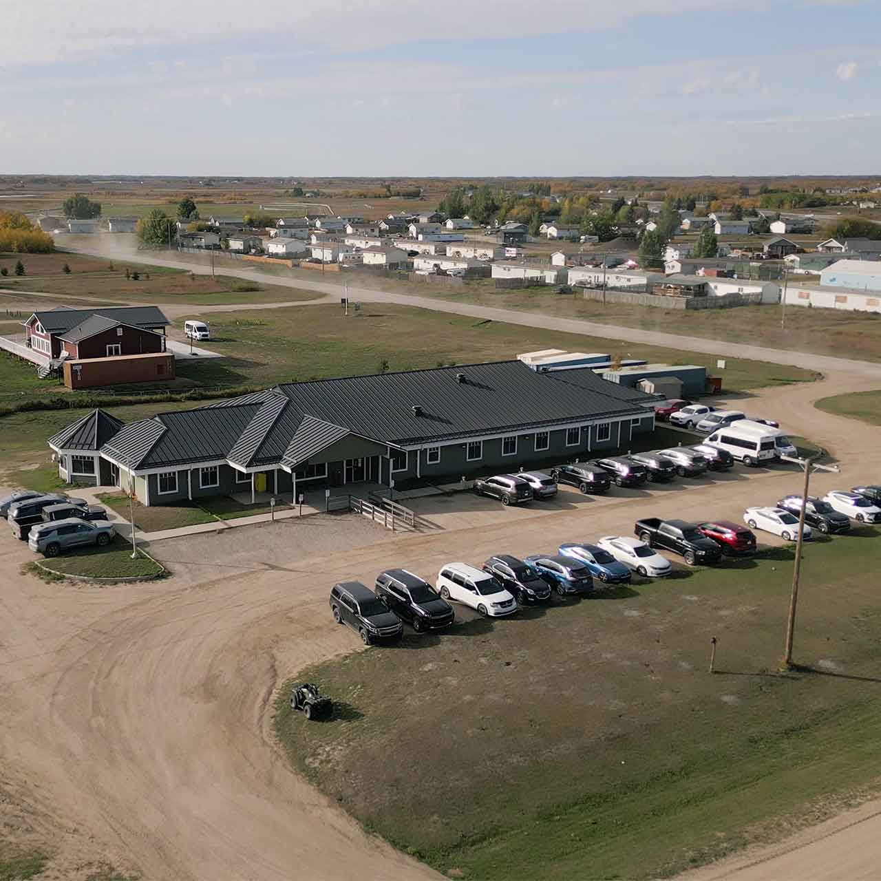 Aerial view of a large single-story building with multiple parked cars on a dirt and grass lot in a rural area with scattered houses in the background.
