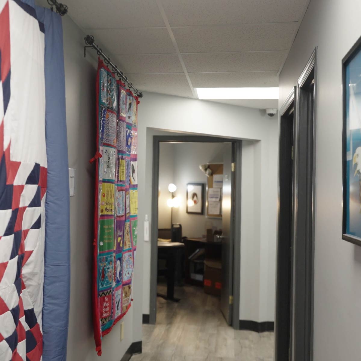 Hallway with handmade quilt hanging on the left wall and an open door revealing a lit office with tables and framed pictures.