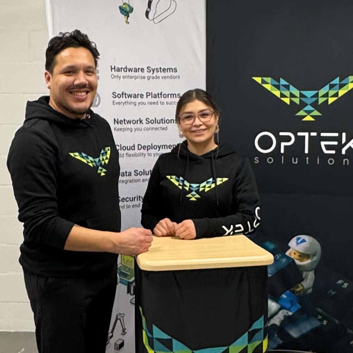 Two people smiling wearing black hoodies with colorful geometric bird logos, standing behind a podium with a matching design and company banners in the background.