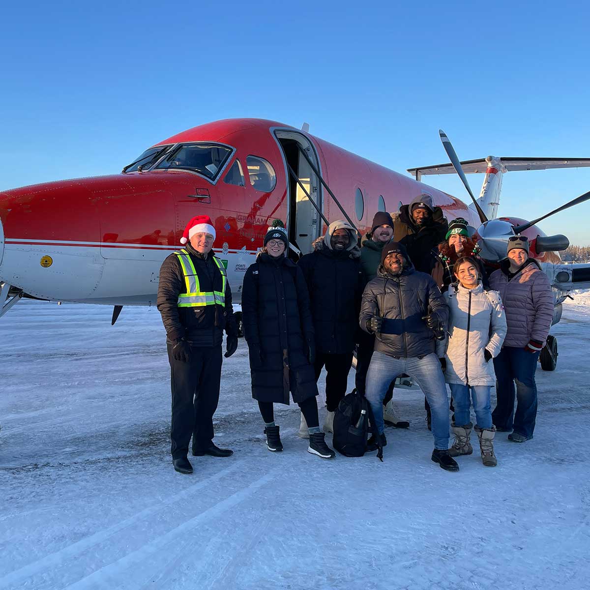 Group of nine people dressed in winter clothes standing in front of a red and white small airplane on a snowy runway, one wearing a Santa hat.