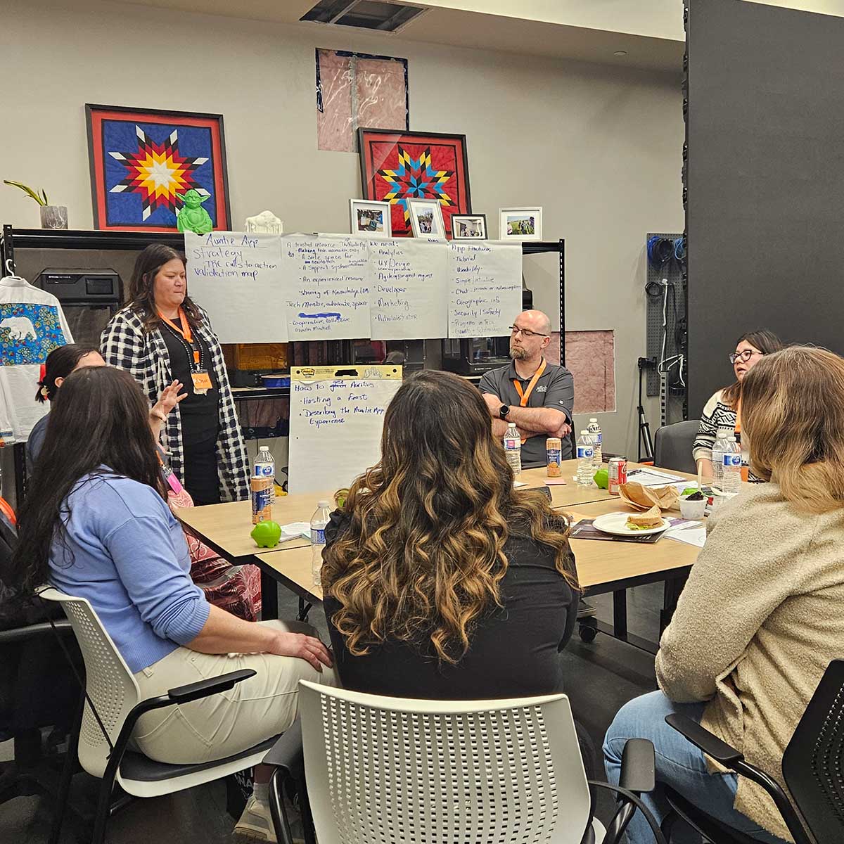 A group of people sitting around a table engaged in a discussion, with notes and water bottles on the table and large sheets of paper with handwritten notes on the wall behind them.