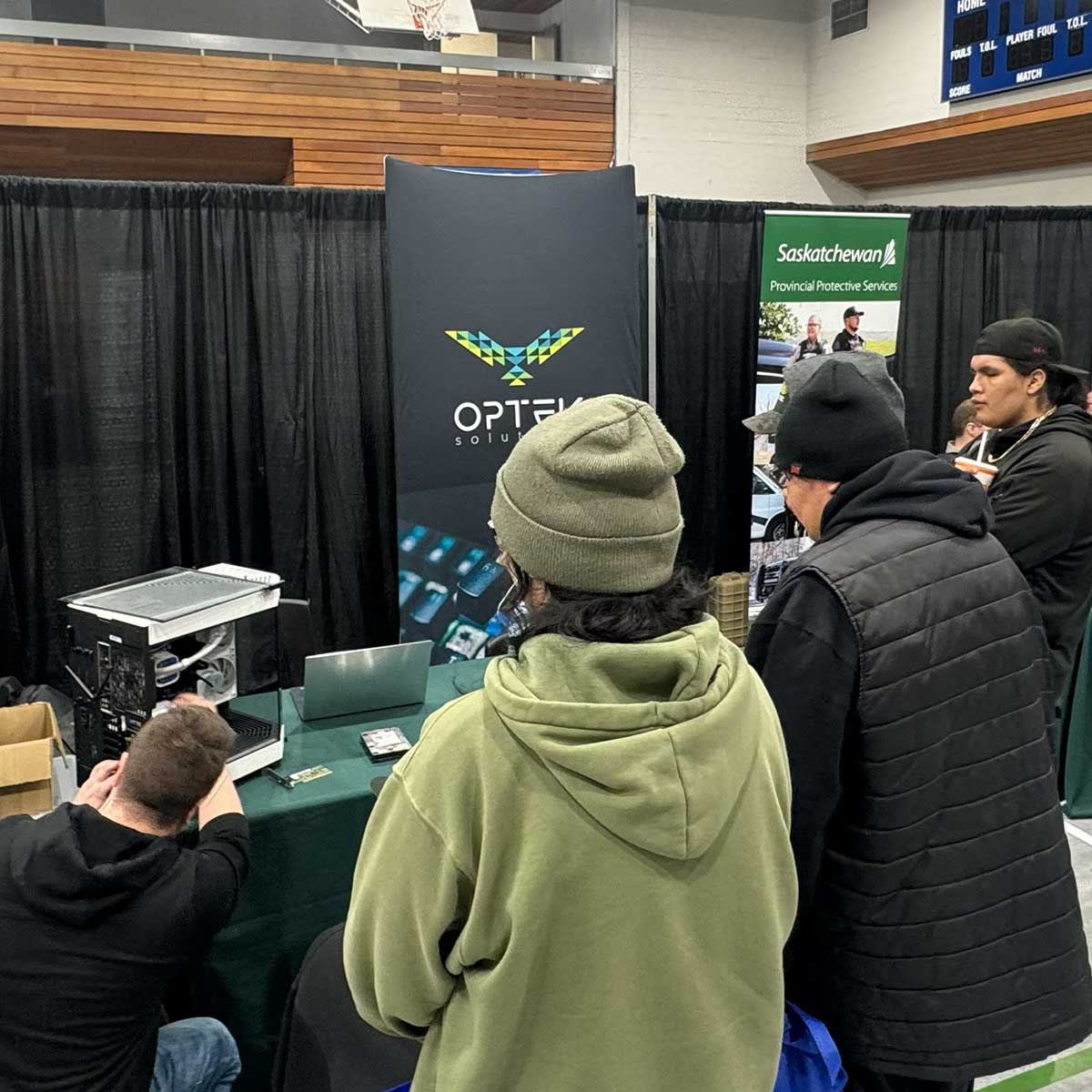 Group of people at an indoor event booth with a black banner displaying the Optera Solutions logo and a Saskatchewan Provincial Protective Services banner in the background.