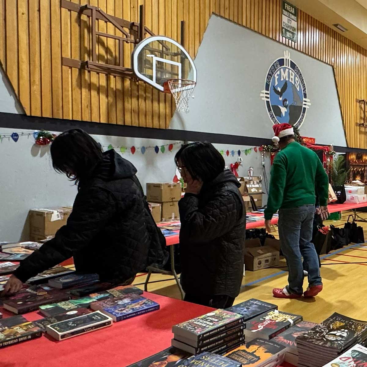 People browsing books on tables in a gym decorated with holiday lights and a basketball hoop.