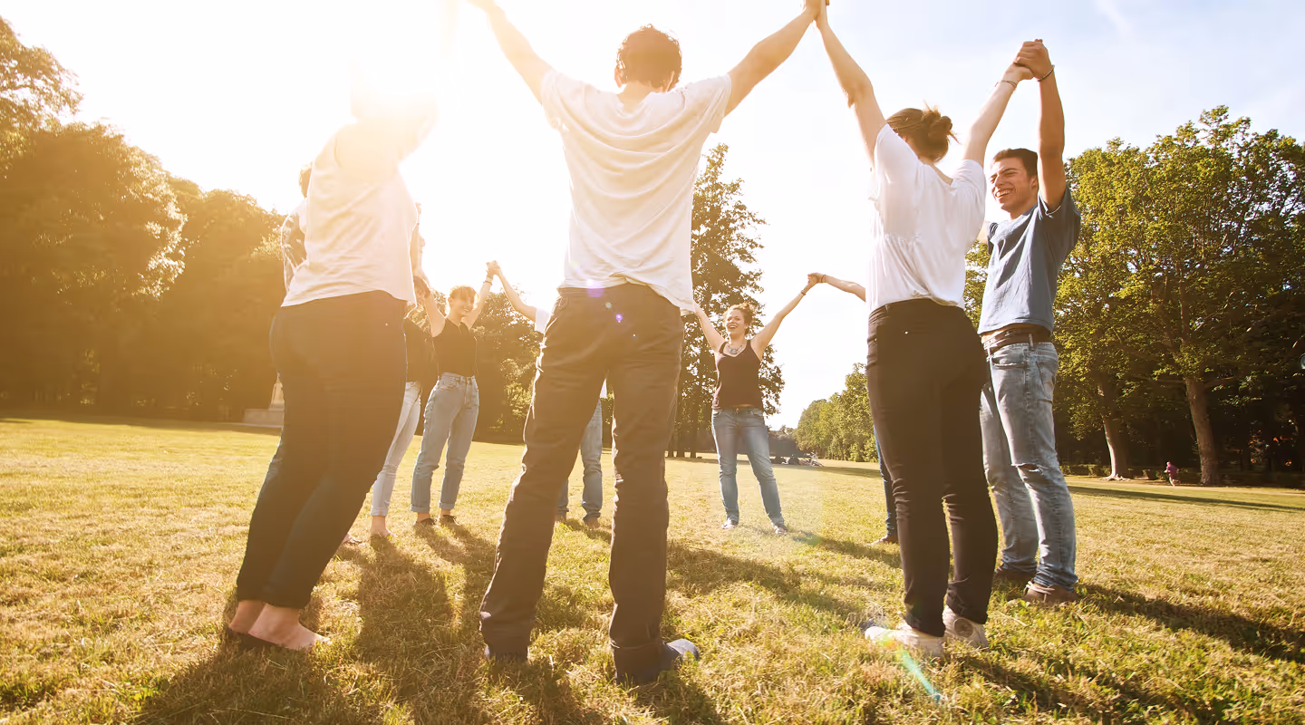 Group of people standing in a circle outdoors holding hands raised under bright sunlight in a park.