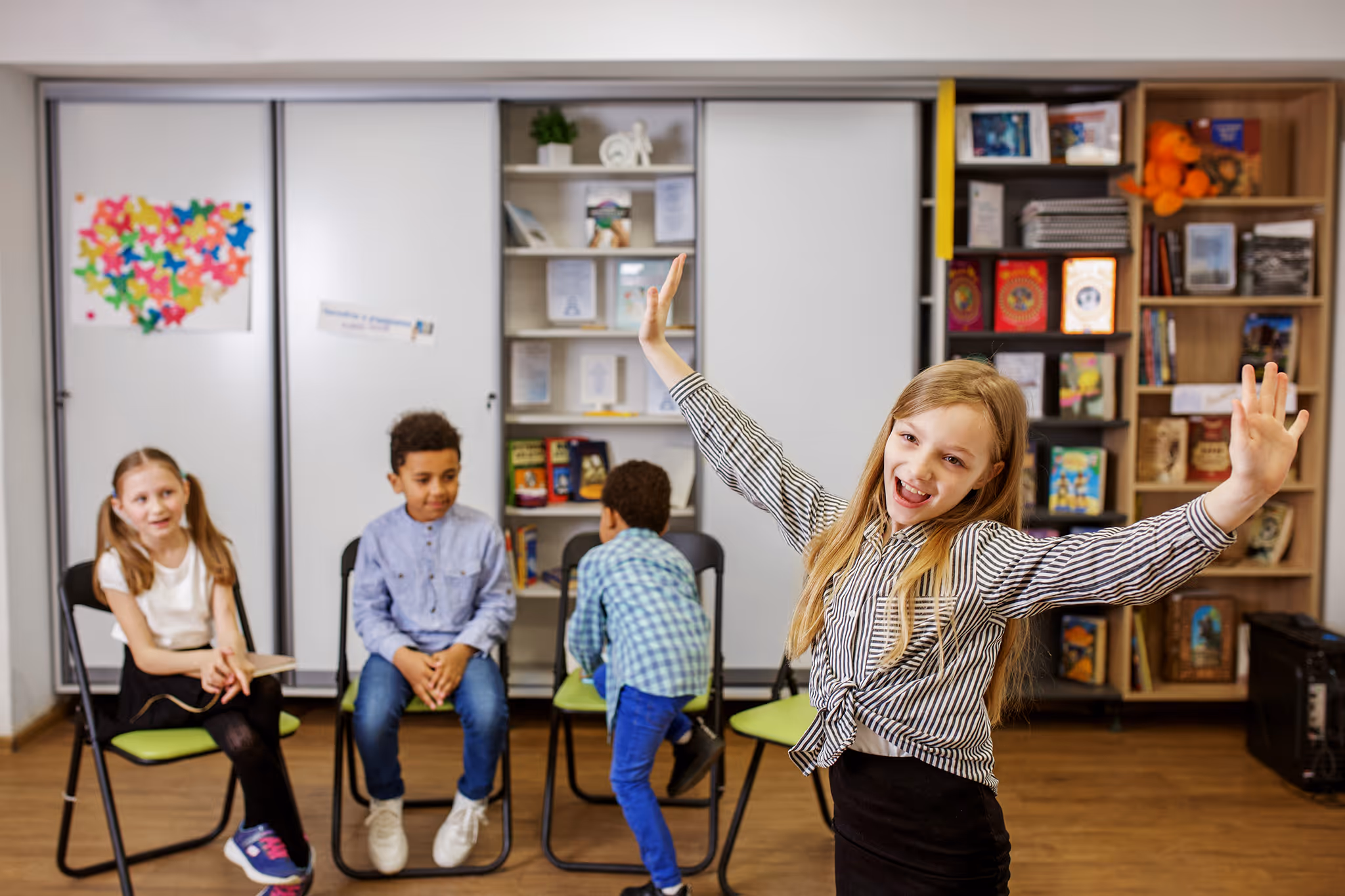Four children in a classroom; one girl stands smiling with arms raised while three kids sit on chairs behind her.