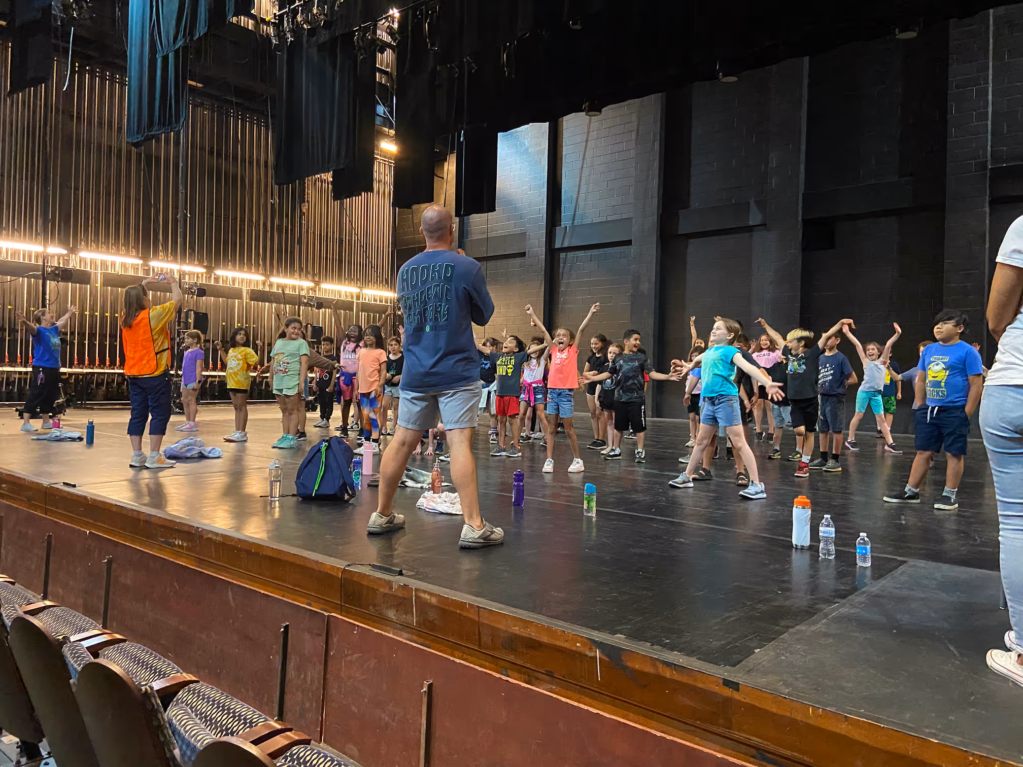 A group of children rehearsing on a stage with instructors, surrounded by water bottles and backpacks.