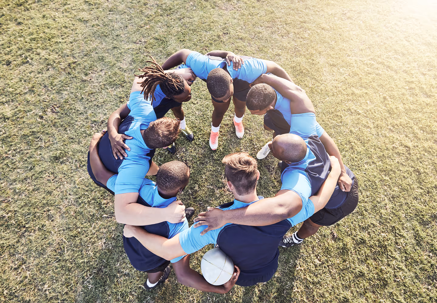 Overhead view of a rugby team huddling together on grass with one player holding a rugby ball.