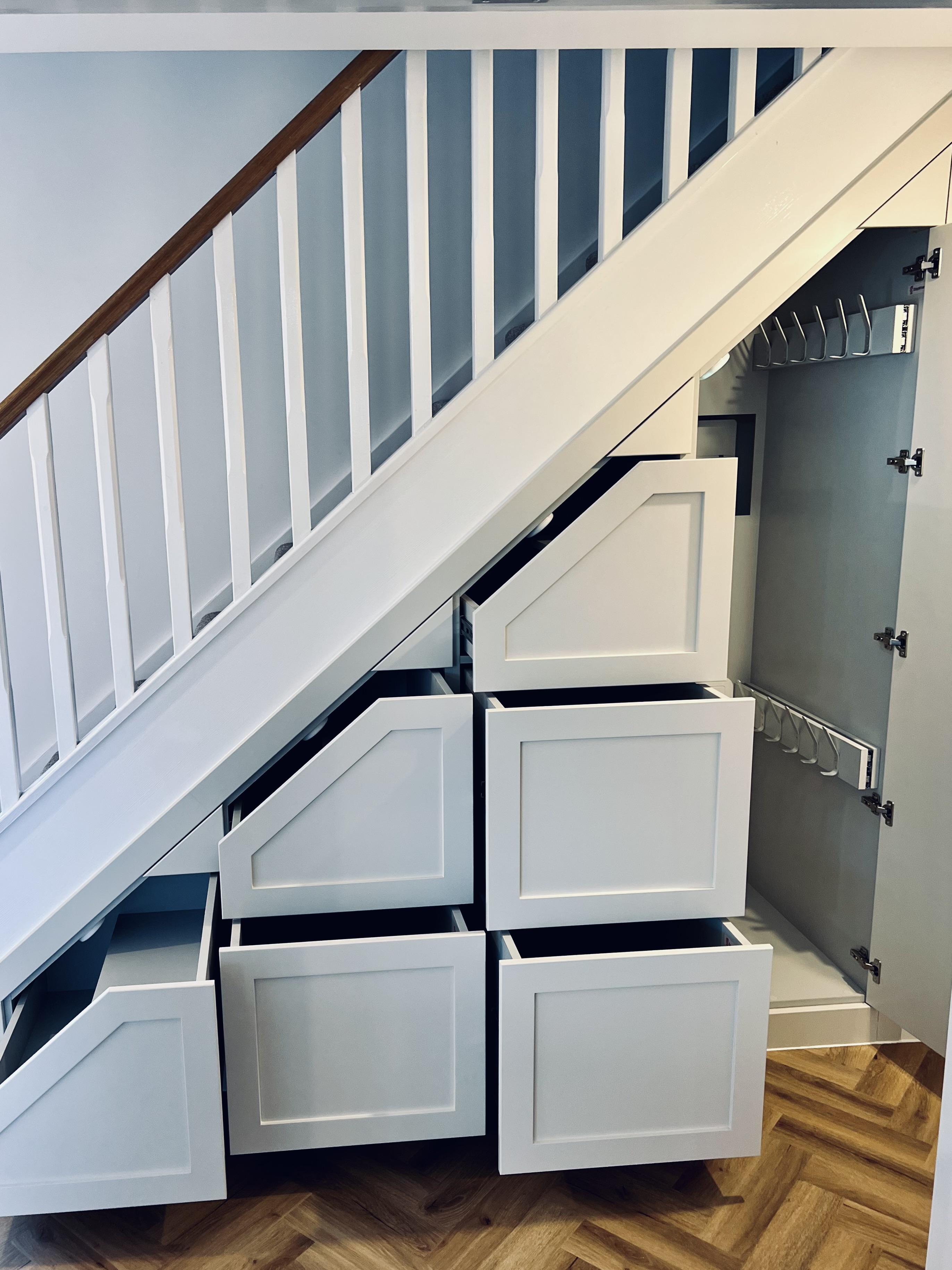 White built-in under-stair storage with open drawers and a cabinet with hooks, on wooden herringbone floor.
