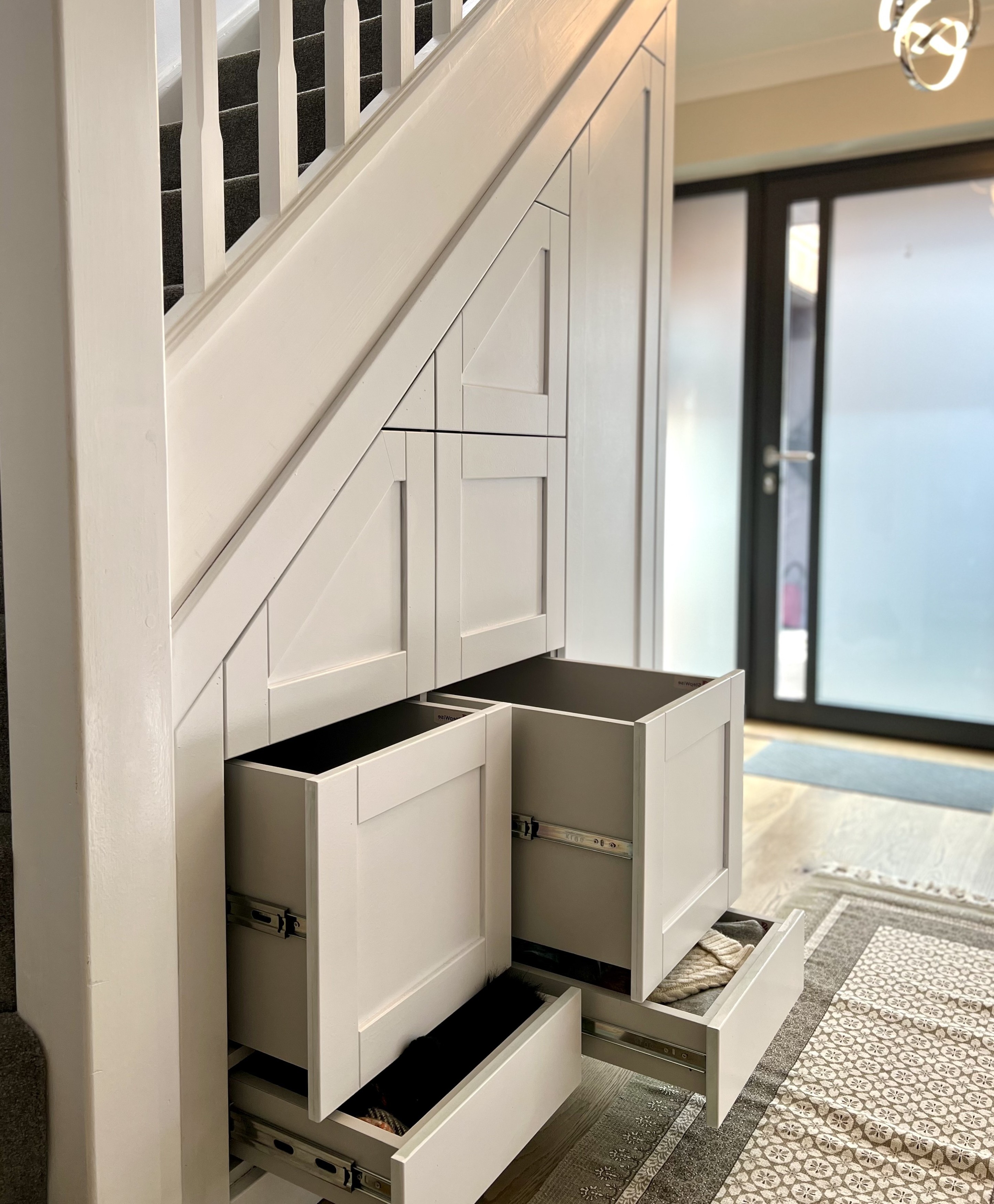 Under-stair white built-in storage with multiple open drawers, next to carpeted stairs and a glass door entrance.