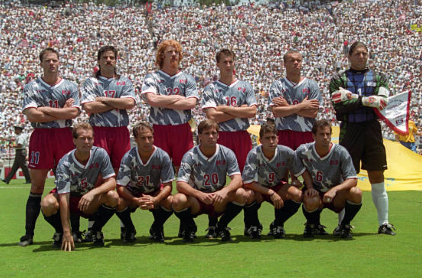 A team photo of USA Men's National Team lining up before a game in the USA 1990 World Cup in the grey, white jerseys and red shorts.