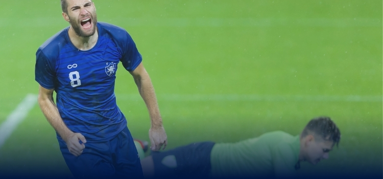 A professional soccer player celebrating a goal with goalkeeper on the ground behind him.