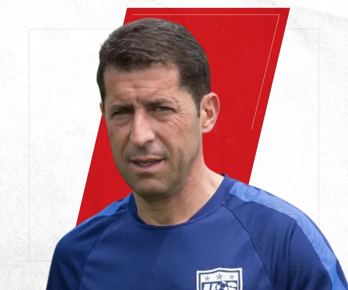 Man with short dark hair wearing a blue US soccer training shirt with a US Soccer crest on the chest in front of a white background with a red shape.