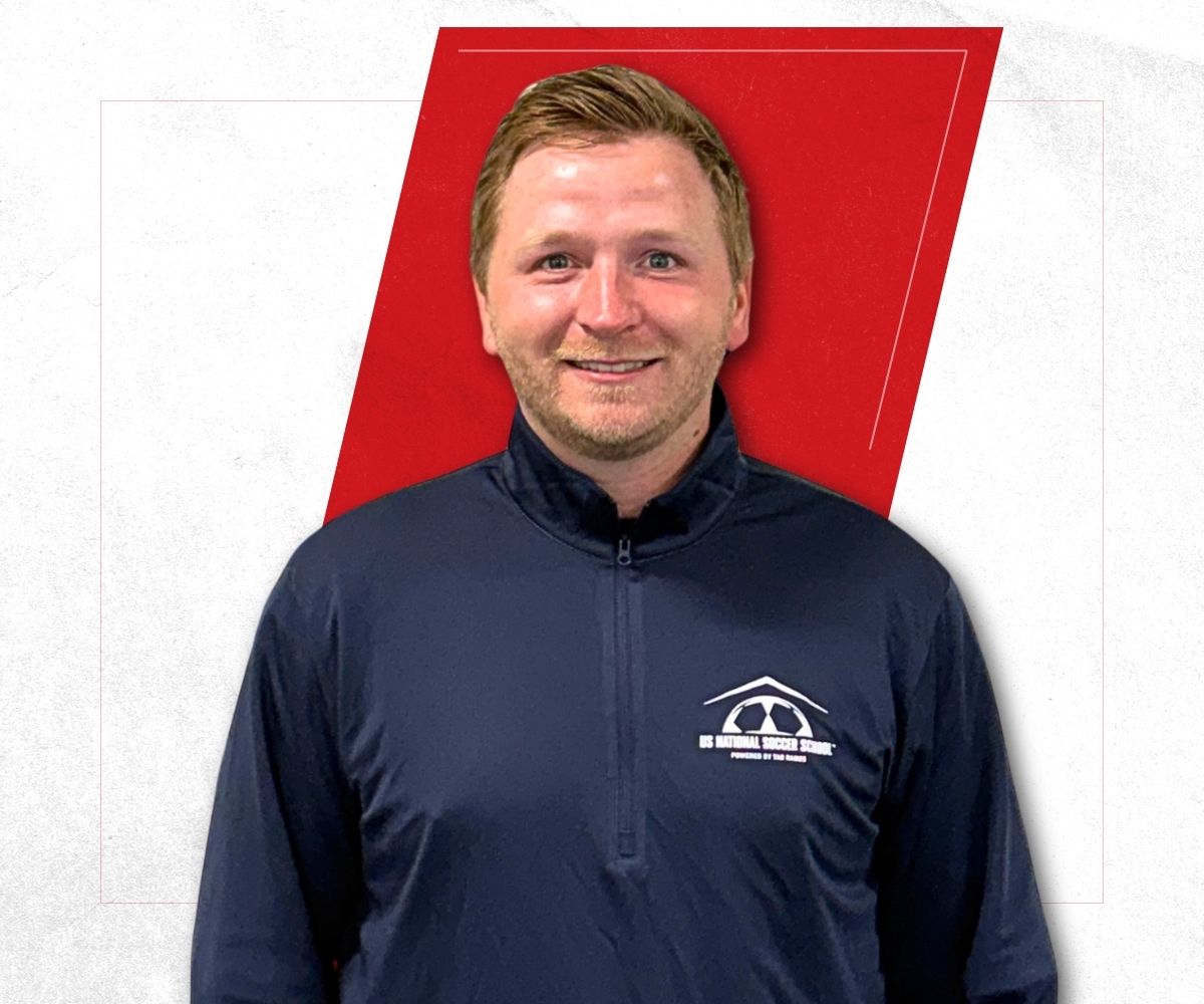 Smiling man with short light brown hair wearing a navy blue quarter-zip jacket with a white US National Soccer School logo on the left chest, standing in front of a white and red geometric background.