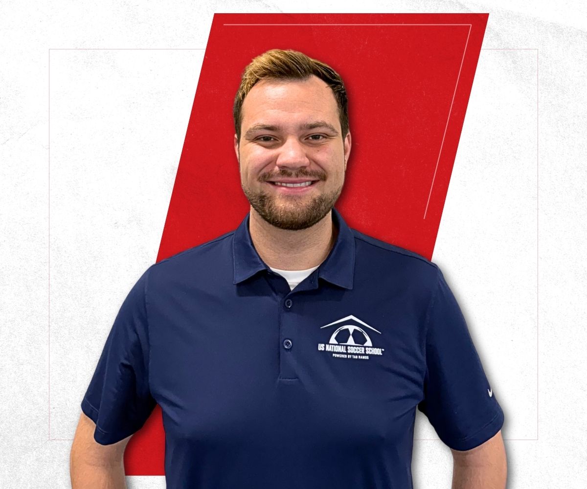 Smiling man with short light brown hair and a beard wearing a navy blue polo shirt with the US National Soccer School logo, standing against a white background with a large red geometric shape behind him.