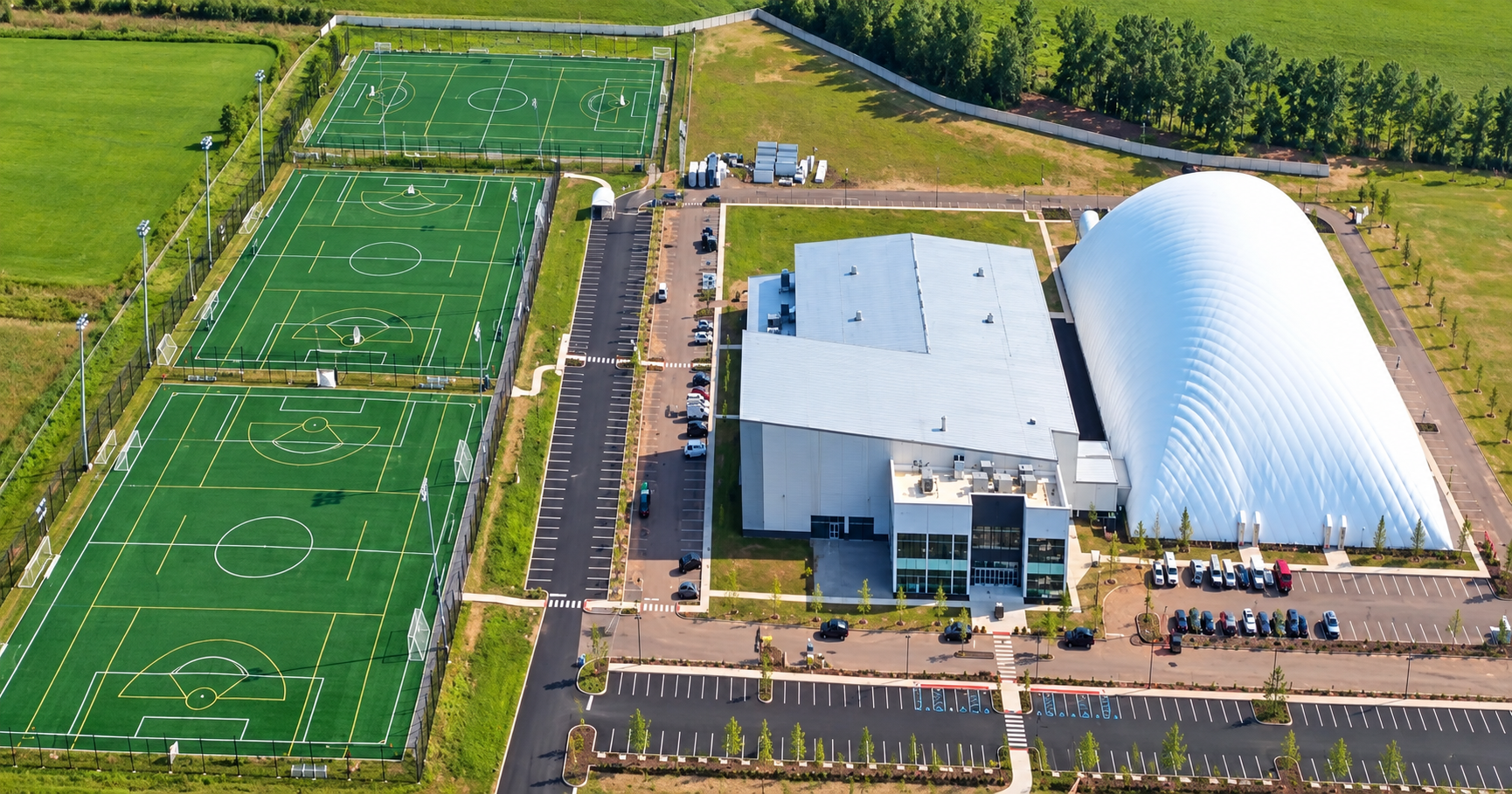 Aerial view of a sports complex featuring three enclosed outdoor soccer fields on the left, a large rectangular building in the middle, and a large white dome structure on the right surrounded by parking lots and green grassy areas.