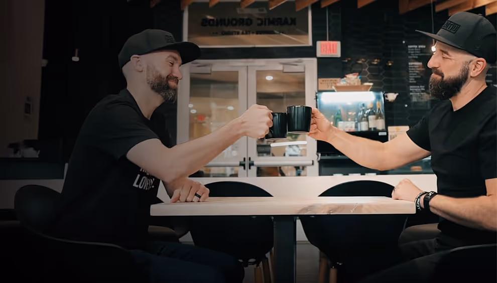 Paul Daly and Kyle Mountsier wearing black shirts and caps sitting at a table and clinking black mugs in a cafe.