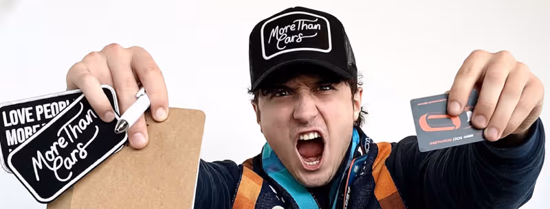 Young man wearing a 'More Than Cars' cap and colorful scarf holds out branded cards and stickers while shouting.