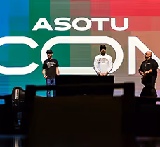Three men standing on stage in front of a colorful ASOTU CON backdrop.