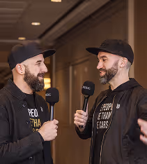 Two men with beards and black caps smiling and holding microphones in an indoor setting.