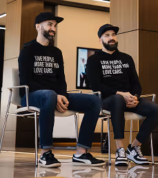Two men with beards and caps sitting on chairs wearing matching black sweatshirts with white text.
