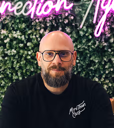 Man with a beard and glasses wearing a black shirt sitting in front of a plant wall with neon text.