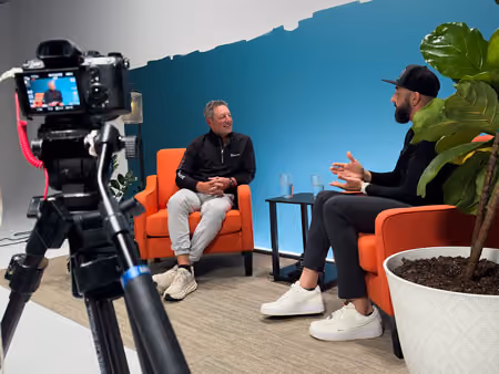 Two men sitting in orange chairs having a conversation while being recorded on camera in a studio.
