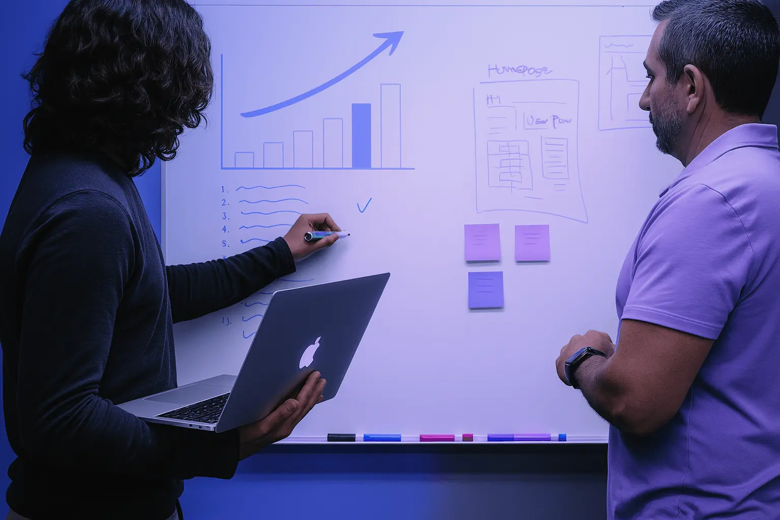 Two people collaborating at a whiteboard with charts, wireframes, and sticky notes, one holding a laptop and writing with a marker.