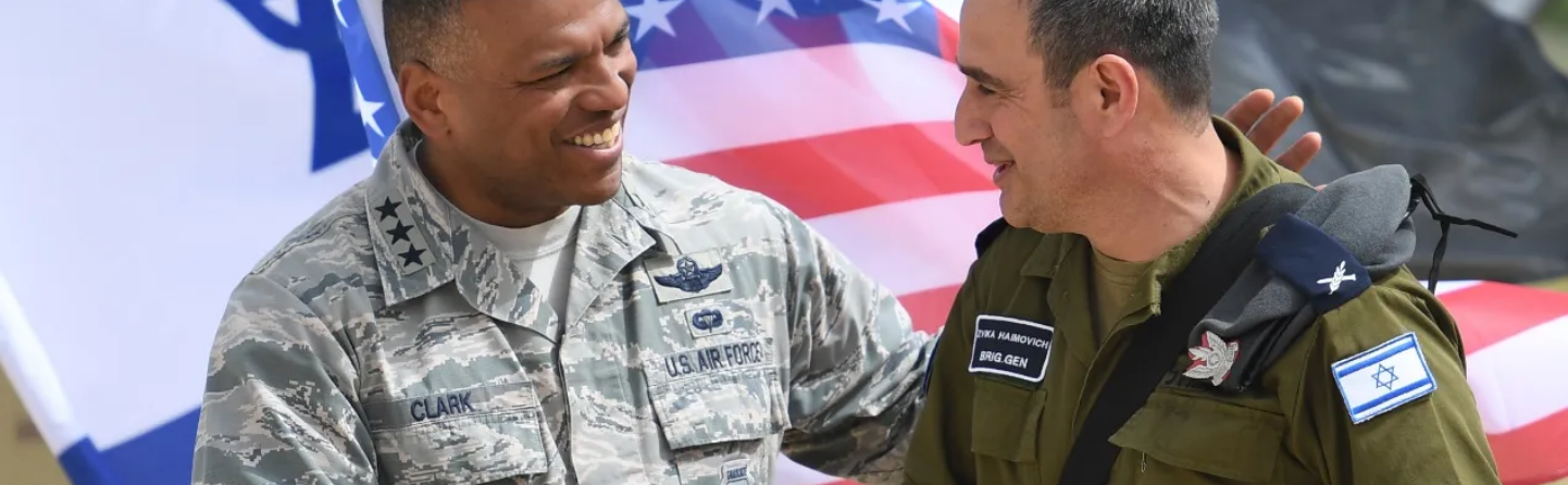 American and Israeli military officers shaking hands in front of American and Israeli flags