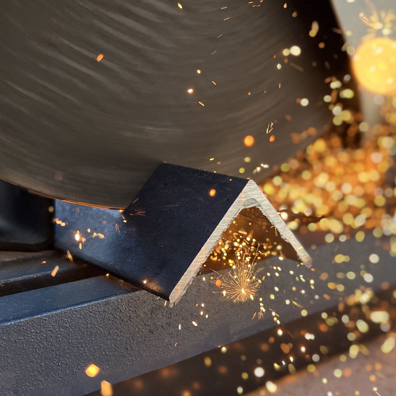 Close-up of a metal cutting saw blade slicing through a black angled metal piece with bright sparks flying around.