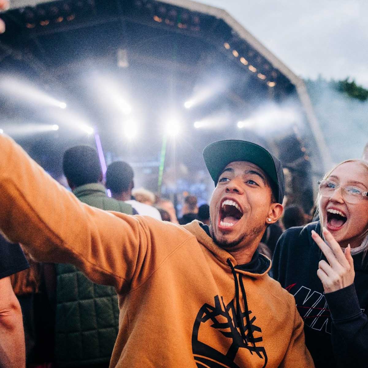 An energetic photo of two people smiling and taking a selfie at a festival with stage lights in the background